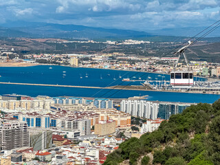 White cable car glides high above the dense cityscape and harbor, offering passengers a panoramic aerial view of the coastline and blue waters from the summit of the famous Rock of Gibraltar.