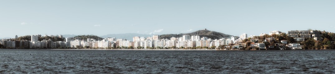 Telephoto panoramic view of Niteroi waterfront skyline, modern apartment towers and hillside houses along Guanabara Bay, Brazil, with calm water, blue sky and distant mountains