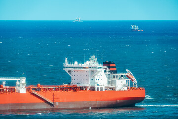Telephoto view of bright orange oil tanker cruising on deep blue sea, white superstructure and deck pipelines, with distant ships on the horizon under clear sky