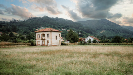 Wide-angle rural Asturias landscape on Camino Primitivo, solitary farmhouse with tile roof in grassy meadow, small village and forested mountains under dramatic sunset clouds