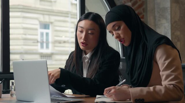 Two diversity business women girls team multiracial females businesswomen at table looking laptop talking Asian boss mentor assistance with corporate data Muslim Islamic intern writing notes in office