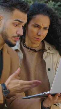 Worried Hispanic couple with laptop confused talking discussing problem in autumn city park on bench family Arabian man with woman looking in computer talk trouble solution difficulty online shopping