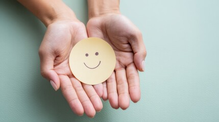 Hands of a person holding a yellow smiley face sticker against a soft green background, symbolizing happiness and positivity in a minimalist setting
