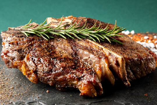 A mouthwatering, juicy grilled T-bone steak, perfectly seared and seasoned with coarse salt, black pepper, and fresh rosemary sprigs, presented on a dark stone surface - Powered by Adobe