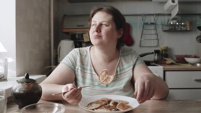 Plus size woman eats tasty pancakes in kitchen at home. High calorie breakfast of baked goods with creamy, sweet sauce leads to obesity. Portrait of person naturally eating food with fork.