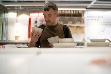 Man in a brown shirt examines ceramic bowls in a store aisle. The setting suggests a retail environment specializing in household goods.