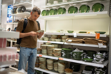 Man with backpack examining a plate in a dinnerware aisle in a store with shelves filled with green and white dishes