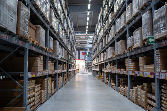 Rows of shelves filled with boxes and pallets in a large industrial warehouse featuring high ceilings and bright lighting.