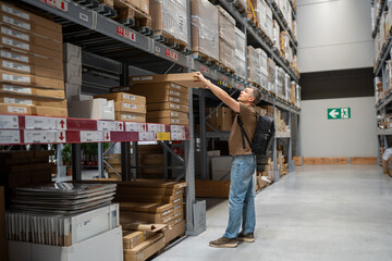 Man wearing backpack reaches for item on high shelf in a spacious warehouse filled with packed boxes and shelves.