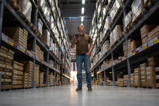 Male worker in a vast warehouse corridor using a mobile device surrounded by tall shelves filled with packages