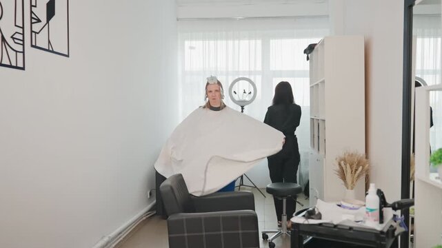 woman in salon cape twirling, stylist prepares tools by mirror and light, minimalist interior with bright window, cape billowing, candid playful mood, professional grooming appointment captured