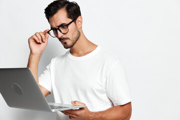 Thoughtful man wearing glasses looking at laptop screen with serious expression, dressed in casual white t-shirt against solid light background. Tech lifestyle concept.