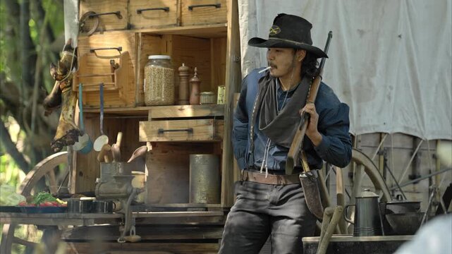 A cowboy stands holding a rifle in a forest camp, with sunlight streaming through the smoke and a chuck wagon in the background