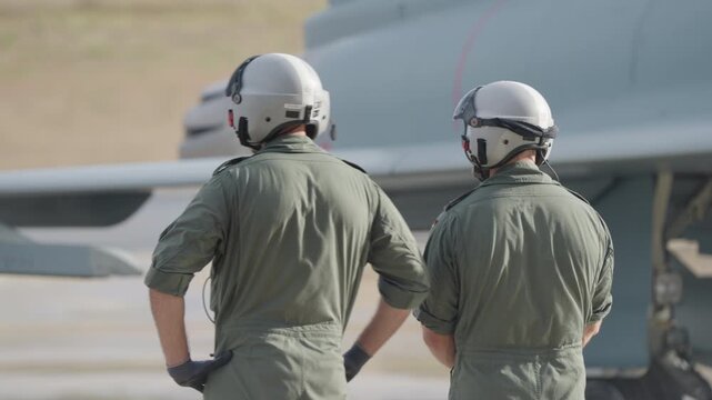 Rear view of two pilots in flight gear observing a fighter aircraft during ground operation on an airbase