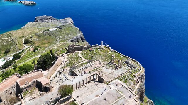Aerial drone video of iconic archaeological site of Acropolis of Lindos built uphill featuring picturesque village, amazing organized beach and Saint Paul bay, Rhodes island, Dodecanese, Greece
