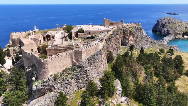 Aerial drone video of iconic archaeological site of Acropolis of Lindos built uphill featuring picturesque village, amazing organized beach and Saint Paul bay, Rhodes island, Dodecanese, Greece