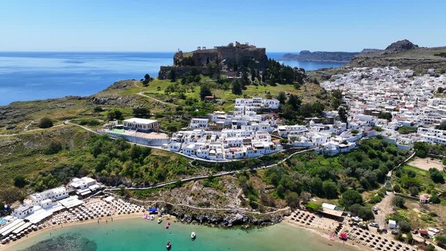 Aerial drone video of iconic archaeological site of Acropolis of Lindos built uphill featuring picturesque village, amazing organized beach and Saint Paul bay, Rhodes island, Dodecanese, Greece