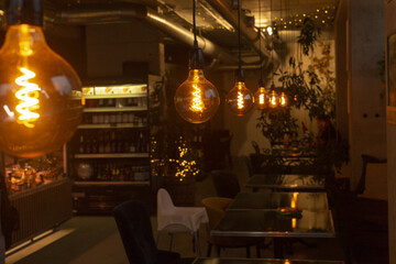 Cozy dimly lit cafe interior with filament bulbs, shelves of bottles, small wooden tables set for...