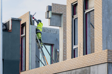 Exterior construction worker performing facade installation on contemporary brick building using ladder, focused on residential construction industry and skilled labor