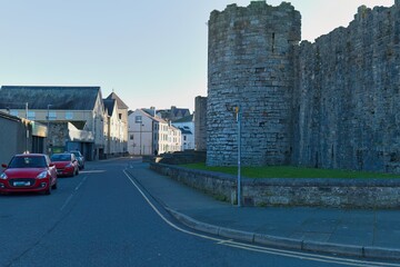 Stone castle wall flanks a street with red cars; buildings line the background under bright sunlight, creating a historic, urban, and slightly serene ambiance in Caernarfon - Gwynedd - Wales - UK