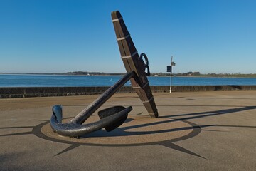 Large black anchor stands angled on a compass rose pavement near blue water under bright, clear sky, casting sharp shadows, creating a nautical ambiance in Caernarfon - Gwynedd - Wales - UK