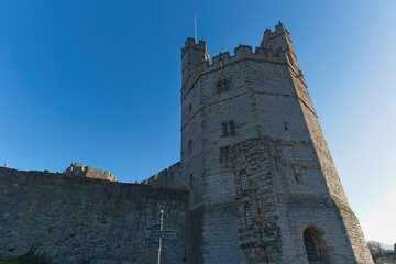 A tall, gray stone castle tower rises against a clear blue sky, connected to a lower, rough-textured wall, bathed in bright, warm sunlight, creating a majestic, historical ambiance in Caernarfon
