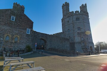 Stone castle gatehouse with turrets connects to a brick building under a clear blue sky, bright daylight, creating a historic, imposing, and solid ambiance in Caernarfon - Gwynedd - Wales - UK