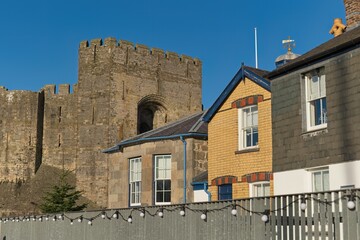 Stone castle wall looms behind colorful houses with white windows, under bright sunlight, creating a historic yet quaint, peaceful, and picturesque scene in Caernarfon - Gwynedd - Wales - UK