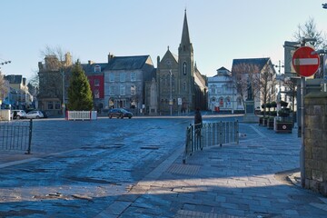 Urban square, stone-paved, bathed in bright sunlight, features a church spire, varied buildings, and a red "no entry" sign, creating a serene, architectural vista in Caernarfon - Gwynedd - Wales - UK