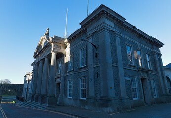 Imposing grey neoclassical building with columns, symmetrical windows, and statues, bathed in bright sunlight, creating a formal, architectural, and slightly austere atmosphere in Caernarfon - Gwynedd