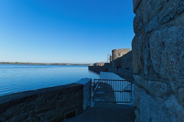 Still, blue water stretches to the horizon under a clear sky, framed by rough stone walls and a metal gate, bathed in bright sunlight in Caernarfon - Gwynedd - Wales - UK
