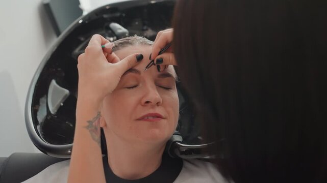 woman receiving eyebrow threading in salon, esthetician with tattooed hands shapes brows over basin, client reclining with closed eyes, precise hand movements, serene expression, warm lighting,