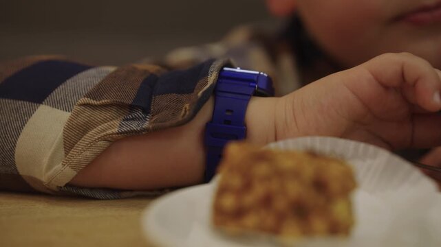 Close-up of child in plaid shirt wearing blue digital watch sitting at wooden table enjoying bite of dessert from small white paper plate, cozy sweet moment indoors. Shooting in slow motion.
