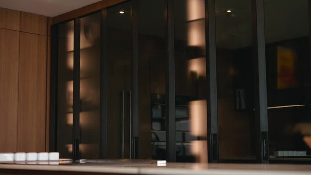 Kitchen interior shows tall dark glass cabinets with reflective surfaces, wooden wall panel behind. Countertop holds small white containers in row and wooden bowl to side