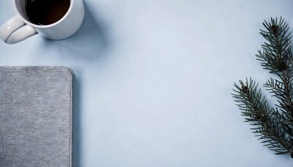 Cozy workspace with a grey notebook, white coffee mug, and pine branch on a light blue surface.