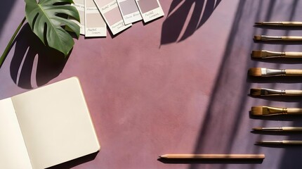 Artist desk setup with paintbrushes, a notebook, color swatches, and a monstera leaf under dramatic window shadows.