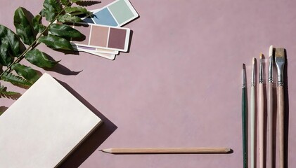 Overhead view of a painting desk with paintbrushes, an open sketchbook, and green leaves on a dusty rose surface.