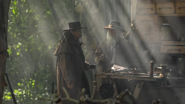 Two senior cowboy stands beside a food cart, drinking coffee while morning light beams through the smoky forest