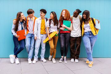 Young happy multiracial student friends standing over isolated blue background. Diverse teenage people having fun talking leaning on campus building college wall. Education and youth concept