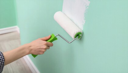 A person's hand holding a green paint roller applying white paint to a mint green wall with a partially painted section.