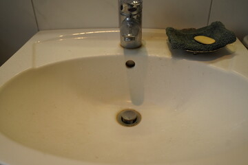 White bathroom sink with chrome faucet, drain, and overflow hole; a green scrub pad and yellow soap sit on the rim. A simple washbasin scene.