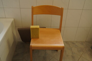 Wooden chair in a tiled bathroom with a yellow sponge on the seat beside a bathtub. Simple indoor scene shows cleaning item on chair