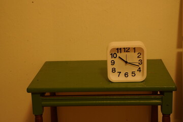 White analog alarm clock on a green wooden table against a beige wall, showing numbers "1 2 3 4 5 6 7 8 9 10 11 12". A simple indoor still life.