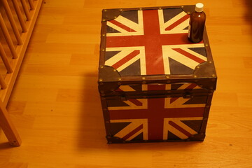 Decorative trunk with a red, white, and blue flag pattern on a wooden floor, topped with a dark bottle. A vintage storage box in a room setting.