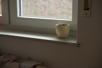 Small ceramic planter on a windowsill in soft daylight, with a blurred view outside and a beige wall below. Quiet interior scene.