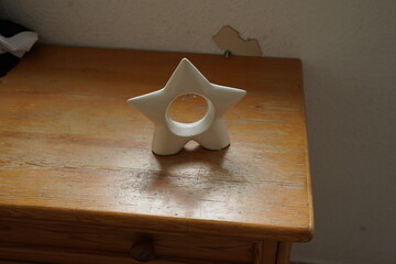 White star-shaped ceramic holder with a circular opening on a wooden table, against a plain wall with a small peeling spot. Minimal still life with a simple star ornament.