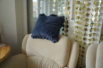 Beige armchair with a blue ruffled pillow near a window, with hanging star garlands in the background. Cozy indoor seating scene.