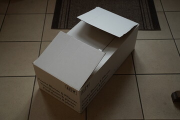 Open white cardboard box with flaps on a tiled floor; small printed text on side is unreadable. An empty package sits on the floor indoors.