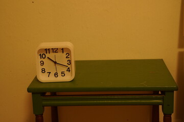 White alarm clock with black numbers on a green table against a plain wall. Simple indoor still life.