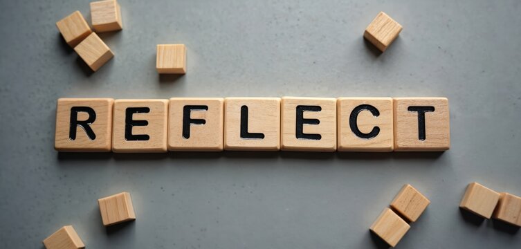 Wooden blocks spell out REFLECT on a gray surface. Scattered cubes surround text. Represents thinking, contemplation, and self-assessment for personal growth.